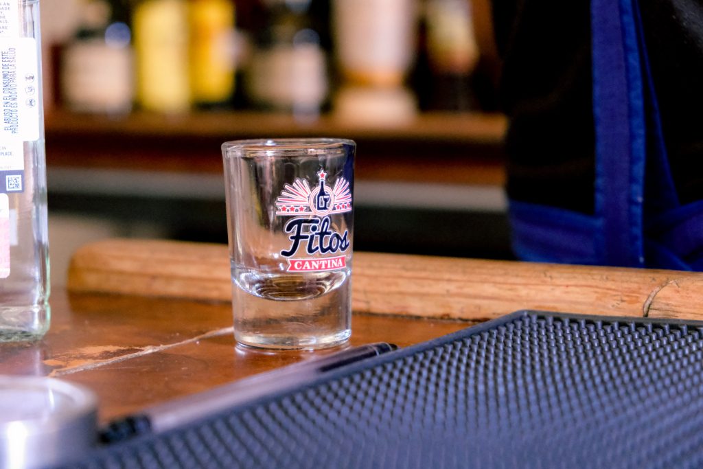 Clear shot glass on a bar counter, labeled Fitos Cantina, with bottles blurred in the background.