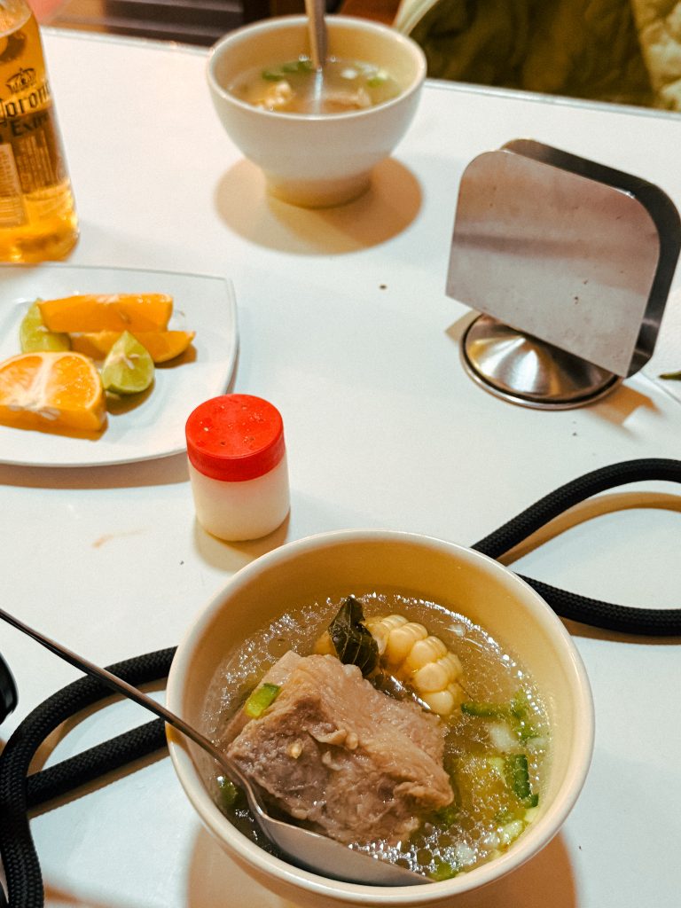 Bowl of soup with meat and corn, alongside a plate of sliced oranges and lime, and a bottle of beer on a white table.