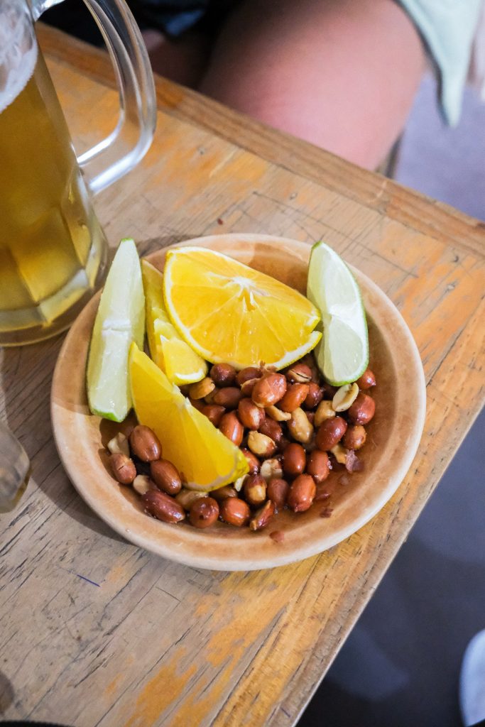 Wooden plate with peanuts, lime, and orange wedges on a table near a beer mug.