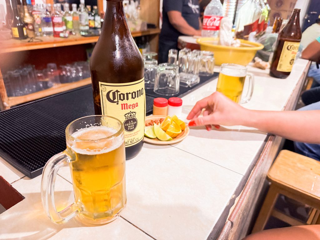 Beer bottle and glass on a bar counter with lemon slices and condiments; a hand reaches for a glass.