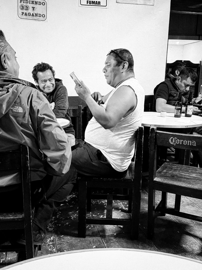 A group of men sitting at a table in a bar, one reading a paper, others talking, with seed strewn across the floor. Black and white photo.
