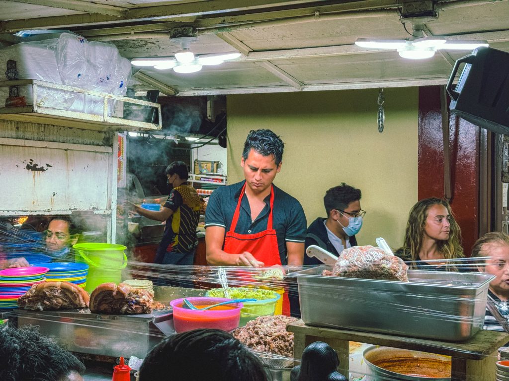 Man in red apron prepares food at a bustling market stall with customers around.