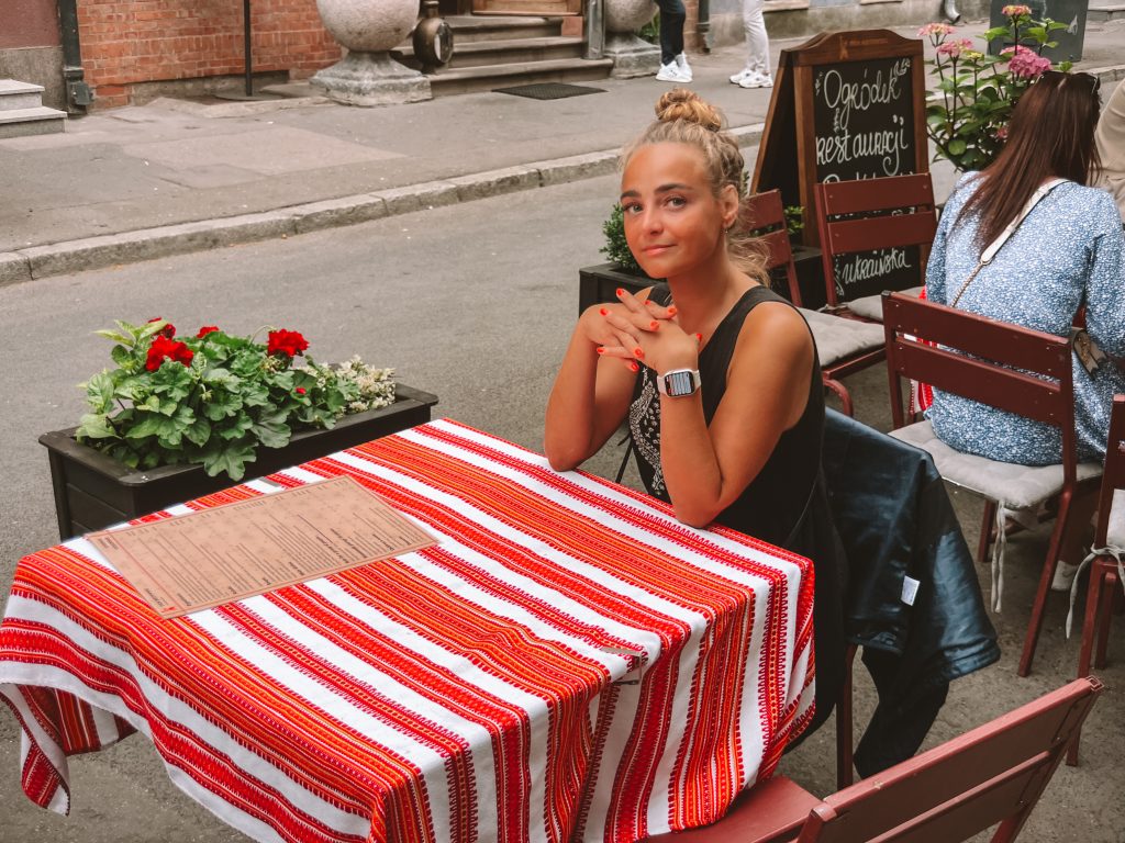 Sofia sitting at an outdoor cafe table with a striped tablecloth, hands clasped, looking at the camera.