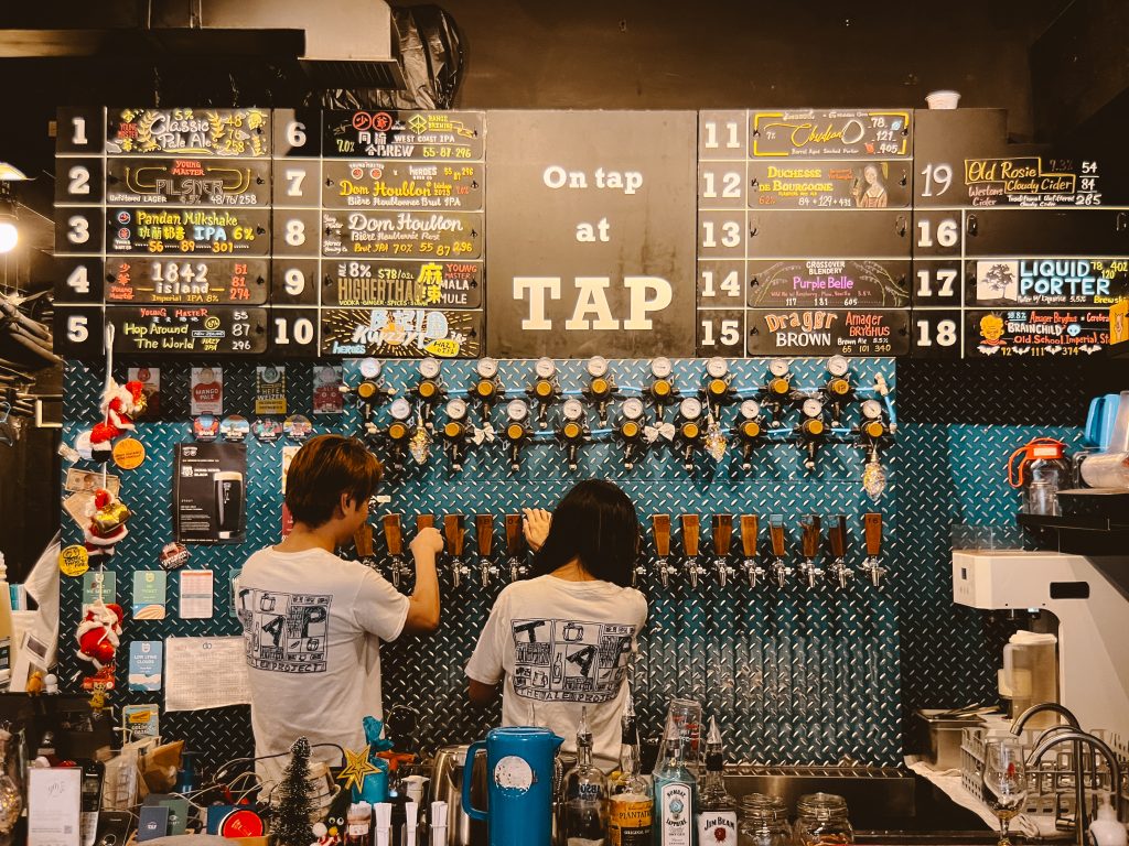 Two people serving drinks from taps in a craft beer bar with a menu board above.