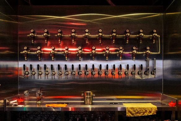 A row of beer taps and gauges on a metallic wall with glasses and a bucket on the counter below.