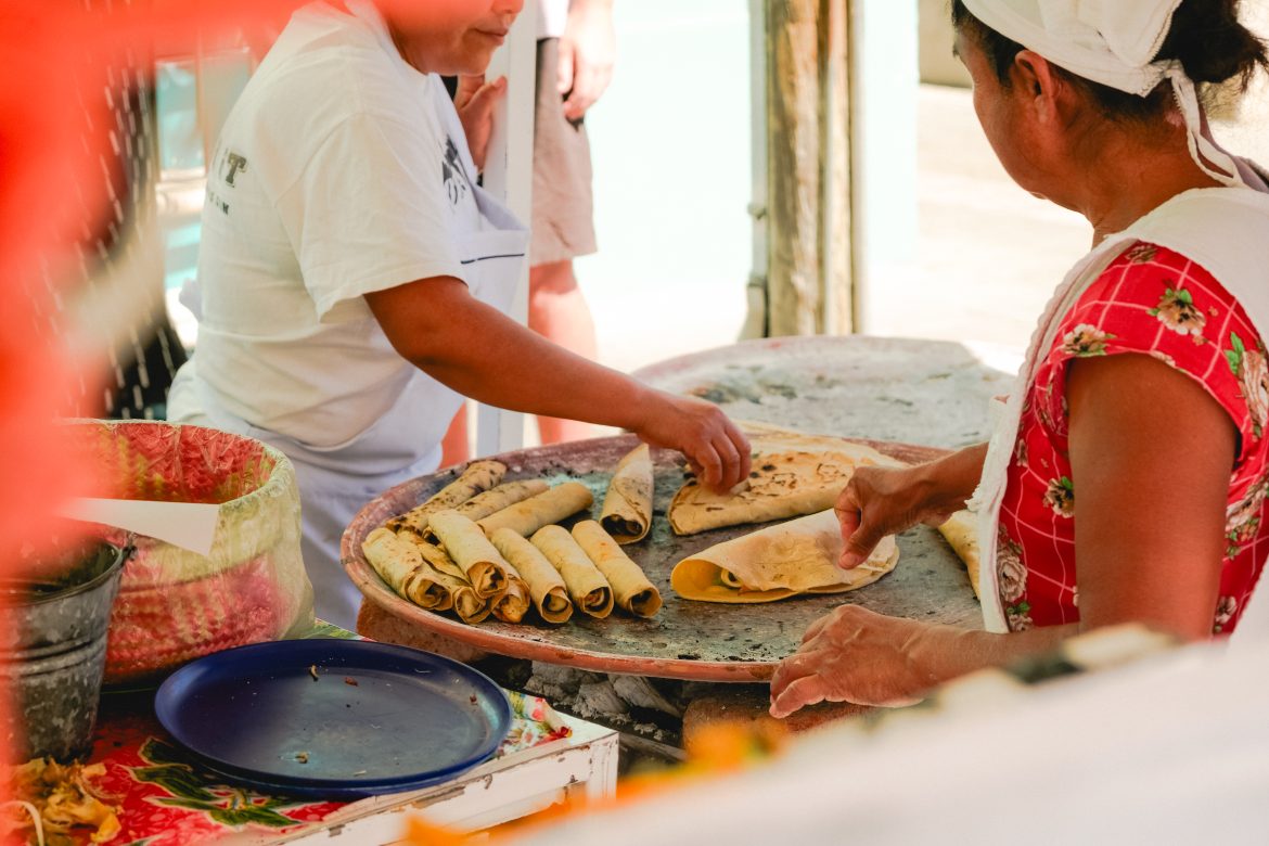 Two people make and arrange tacos on a grill at an outdoor food stall.