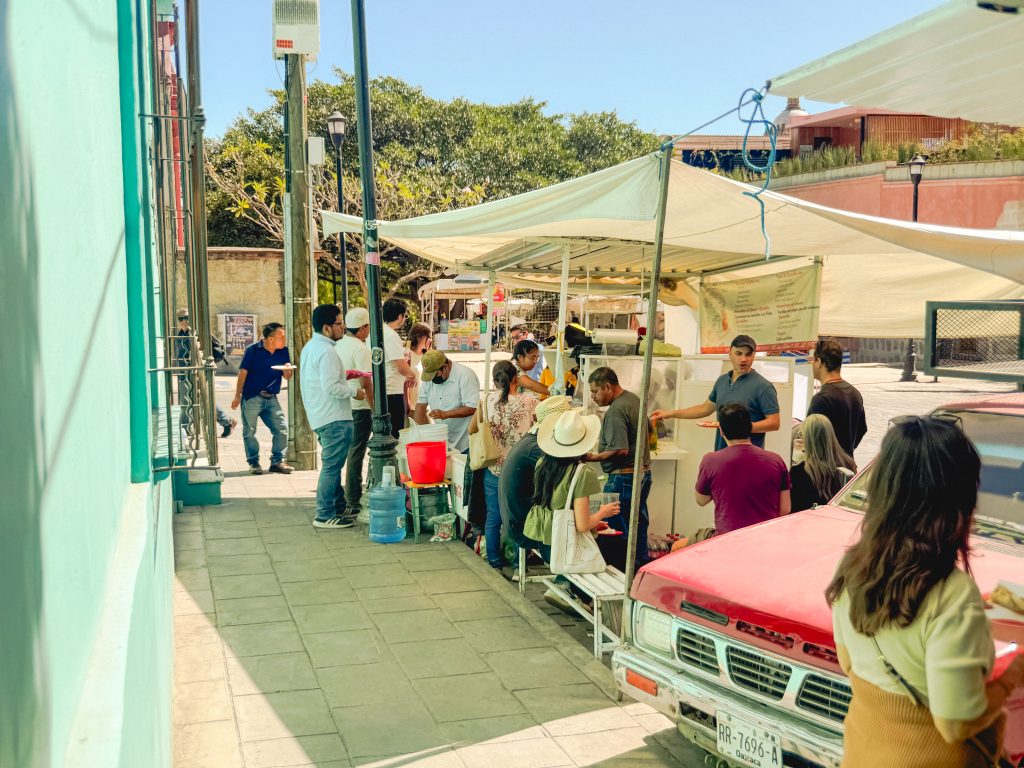 People gather around a street food stall with a red truck nearby on a sunny day in Oaxaca.