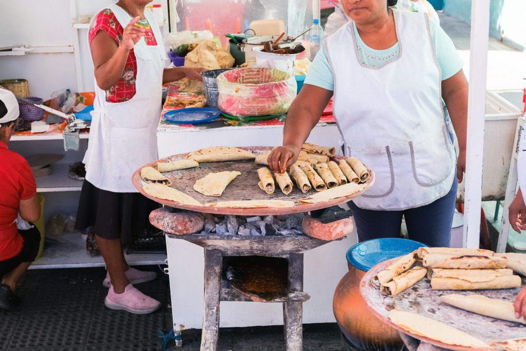 Two women prepare and cook food at an outdoor market stall, with rolled items on a large stove.