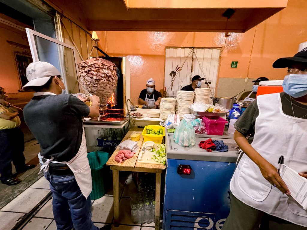 Street food stall with cooks preparing tacos al pastor, stacks of plates, and ingredients on the counter.