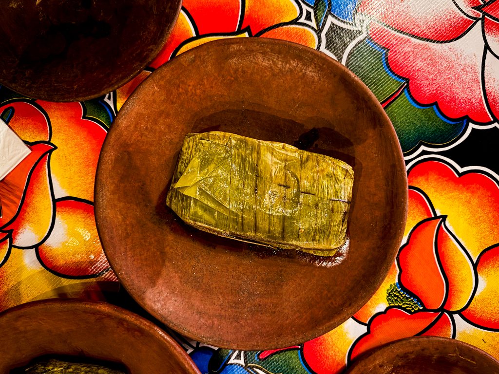Tamale wrapped in a banana leaf on a brown plate, placed on a colorful floral tablecloth.