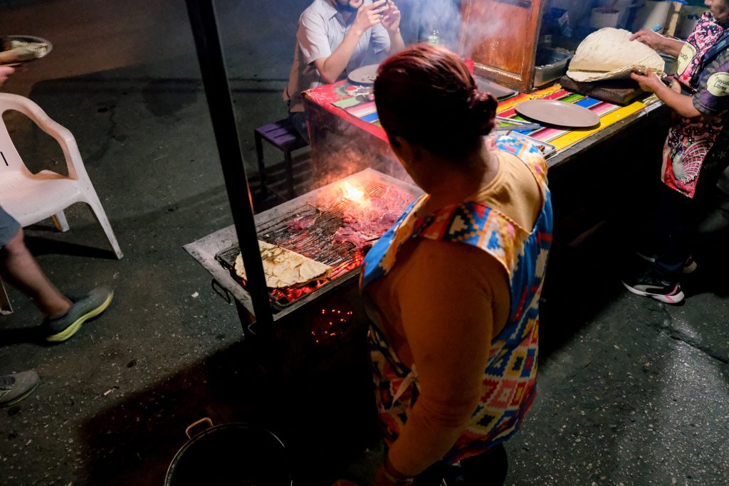 Person grilling food outdoors at night, with others nearby. Colorful tablecloth and plates in the background.