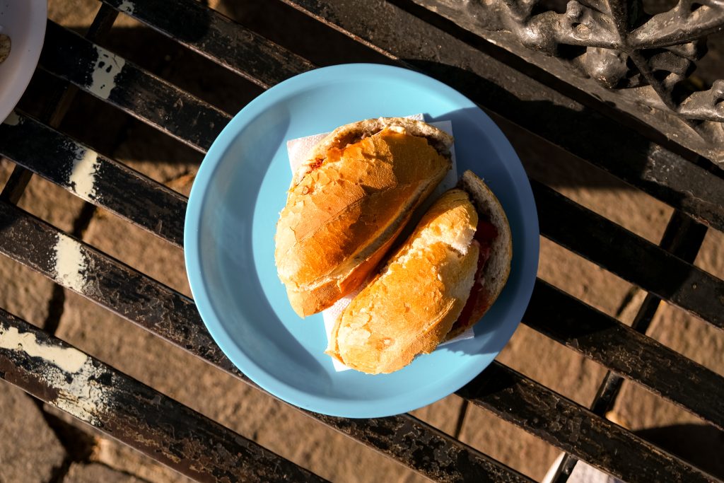 Two sandwiches on a blue plate are placed on a weathered bench in sunlight.