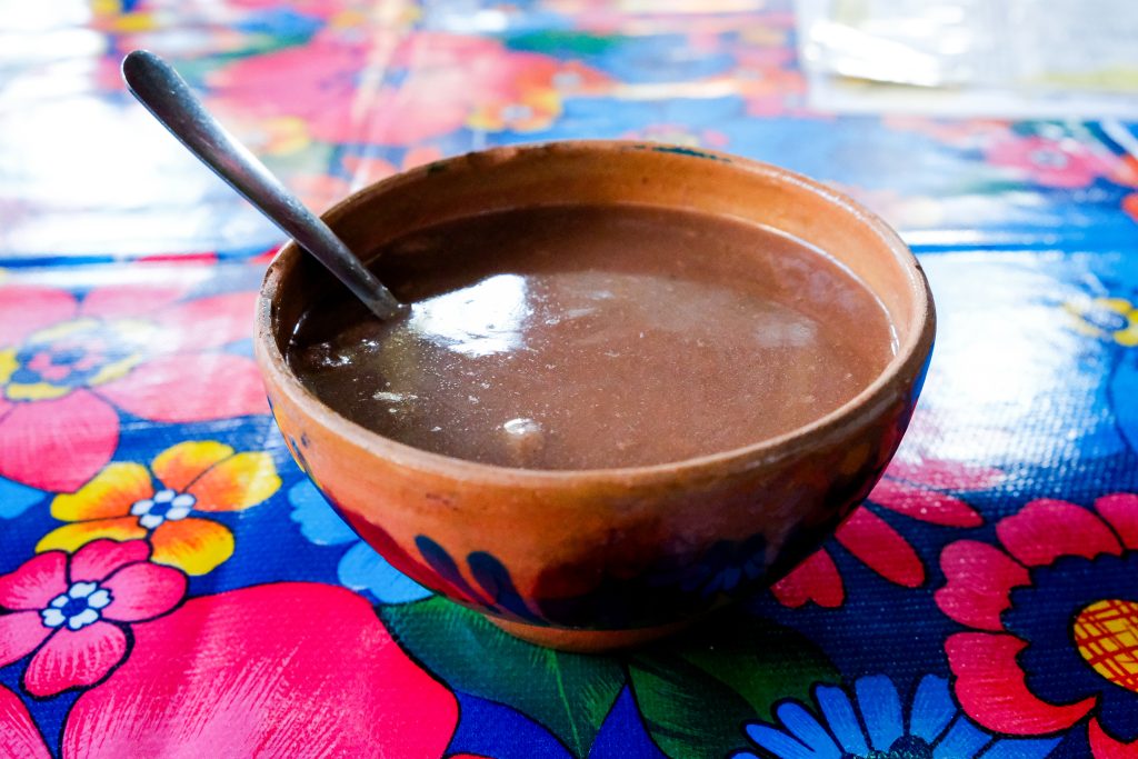 A thick, smooth dark brown chocolate drink sits in a terracotta bowl on a colourful tablecloth at a restaurant.