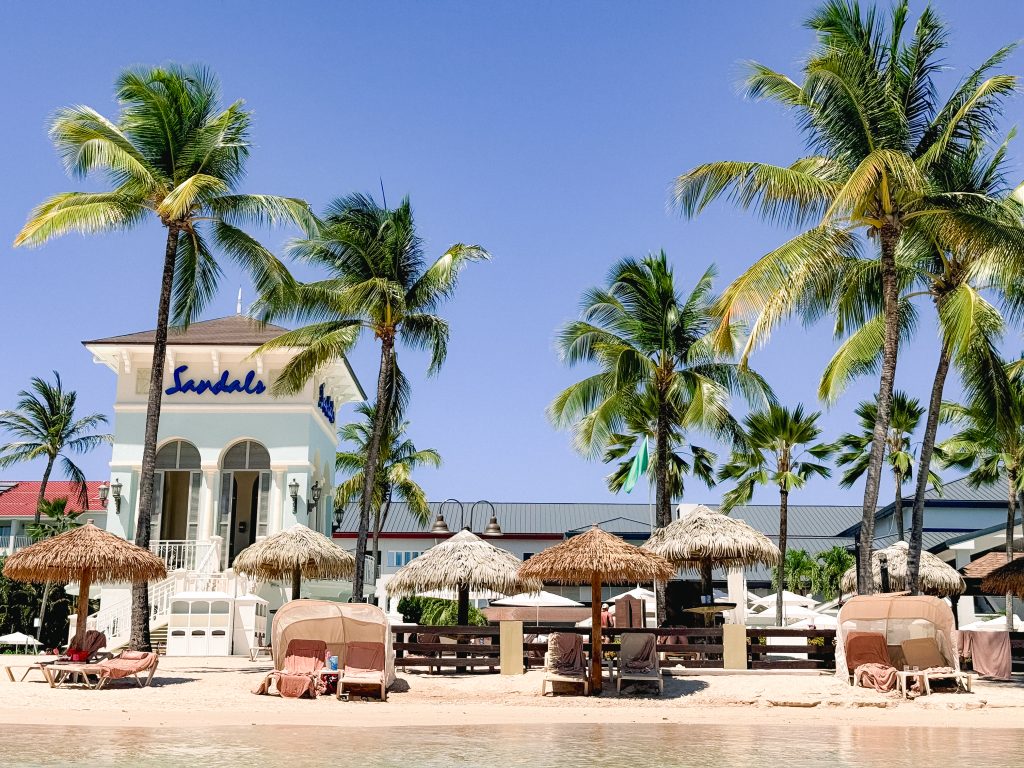 Resort beachfront with palm trees, cabanas, and a building labeled Sandals under a clear blue sky.