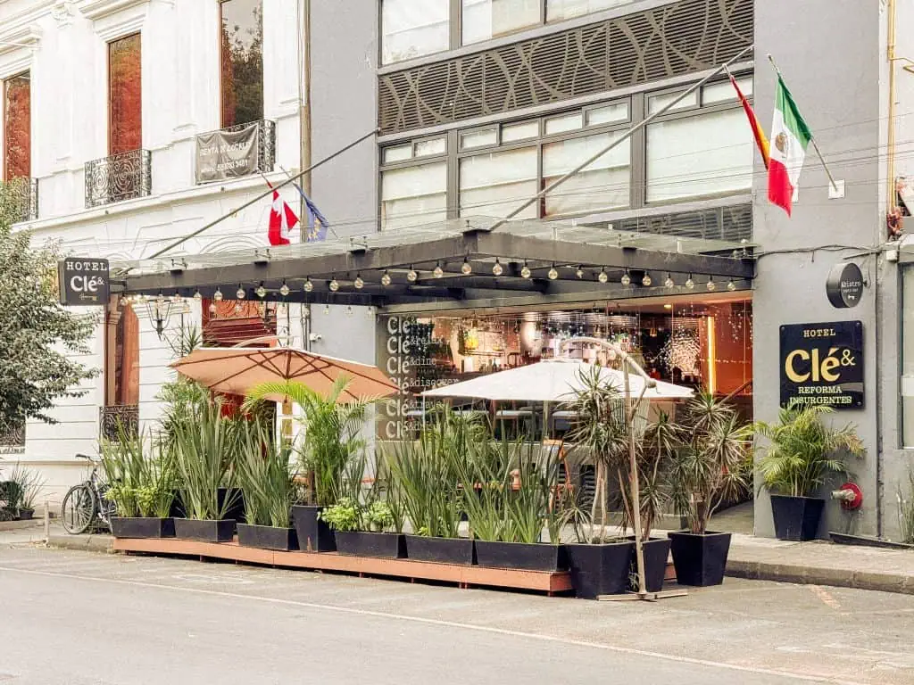 Hotel Clé exterior with flags, potted plants, patio umbrellas, and string lights on a city street.