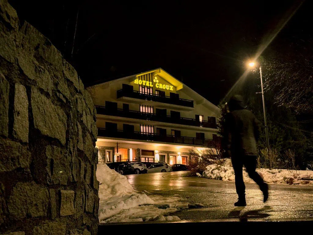 A person walks at night near the illuminated Hotel Croux with snow on the ground.