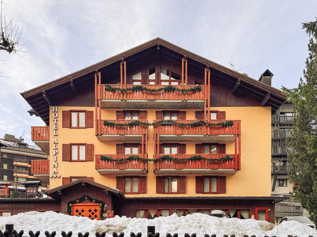 A chalet-style hotel with red balconies and snow on the ground, labeled “Hotel Alpina.”.
