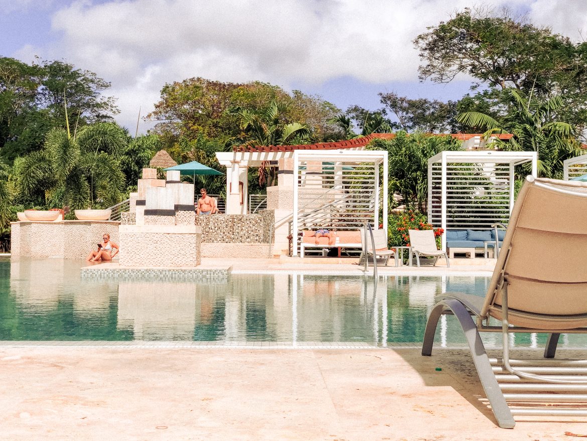 Resort pool with lounge chairs, cabanas, palm trees, and a few people relaxing under the sun.