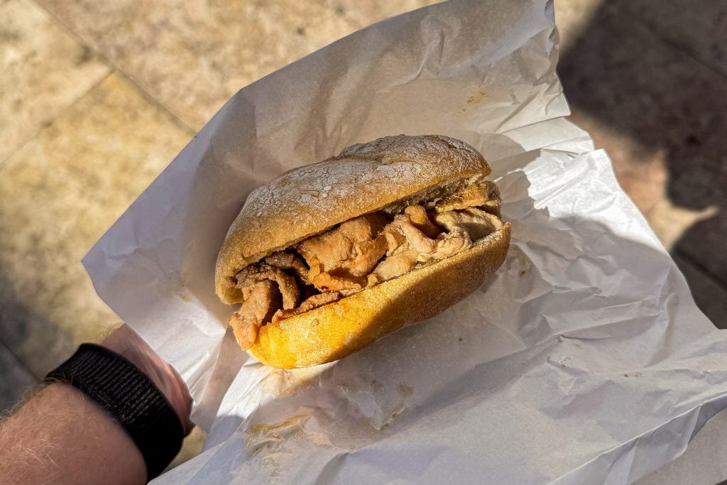 A hand holds a sandwich with sliced meat in a crusty roll, wrapped in white paper.