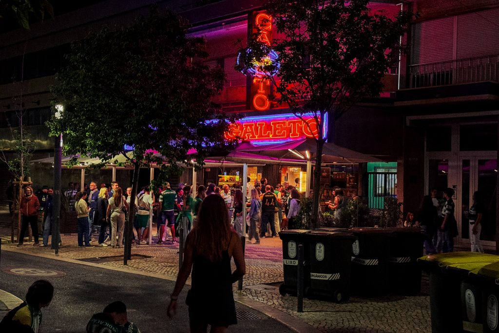 A crowd stands outside a neon-lit bar at night as a woman walks toward the entrance.