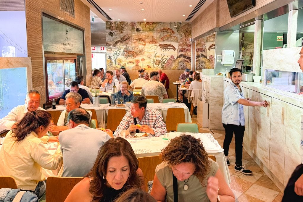 Busy Lisbon restaurant with diners at tables, colorful mural on wall, and a man standing near the kitchen area.