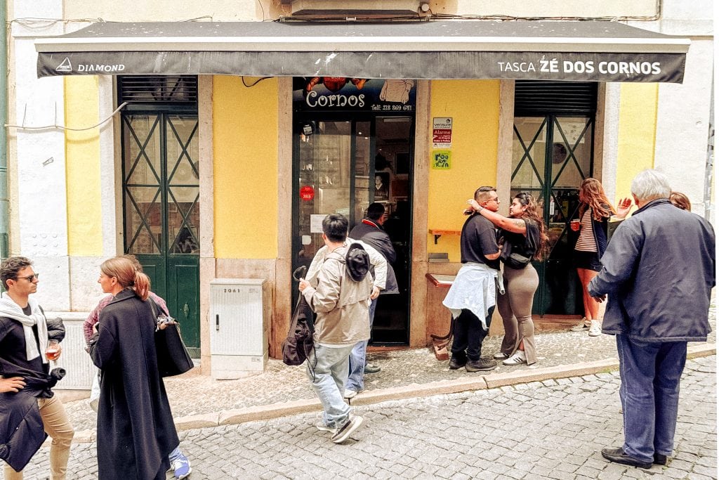 People gather and socialize outside one of Lisbon's best restaurants called Tasca Zé dos Cornos on a cobblestone street.
