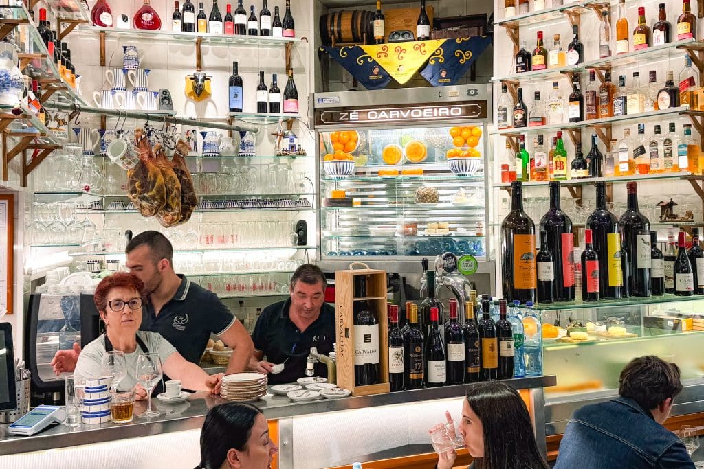 Busy bar with shelves of bottles, glasses, and fruit; staff serving drinks and patrons sitting at the counter.