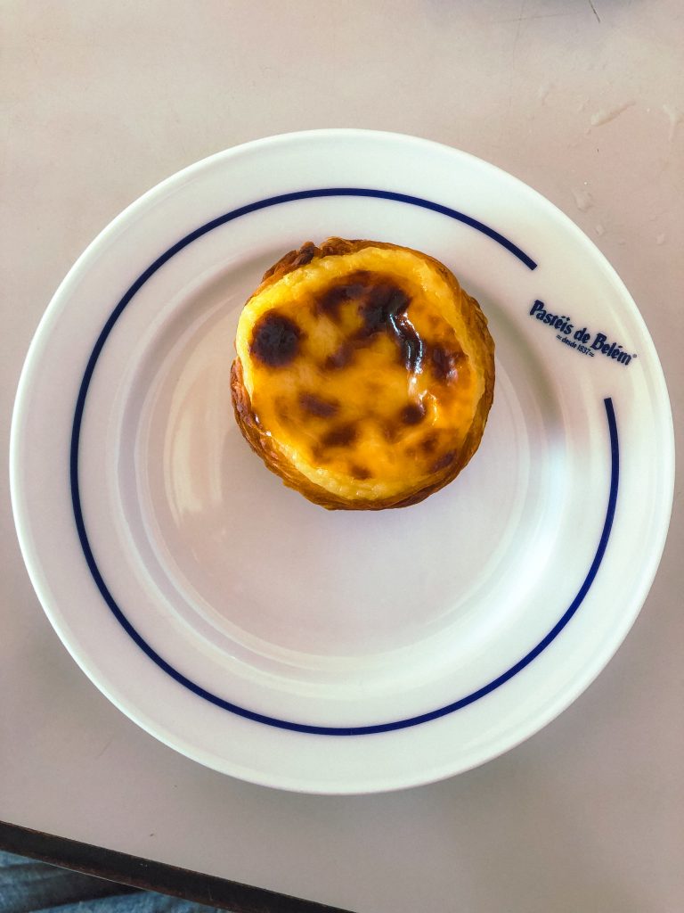 A custard tart on a white plate with blue lines and Pastéis de Belém written on it.