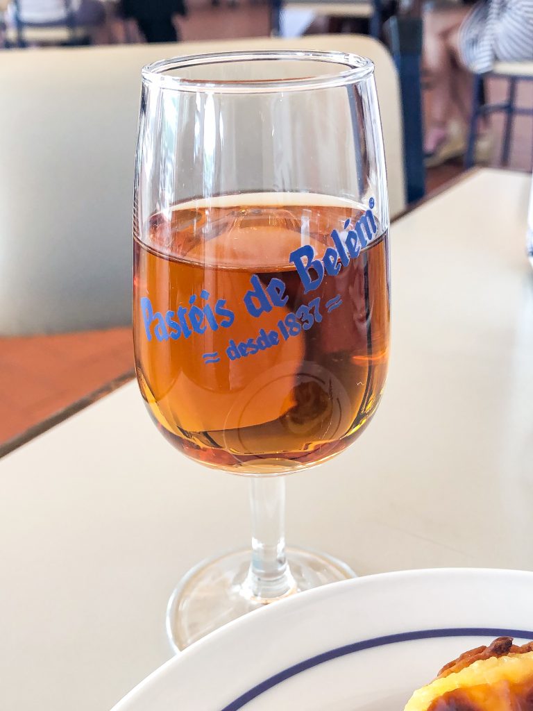 A glass of amber-colored drink with Pastéis de Belém written on it, on a café table.