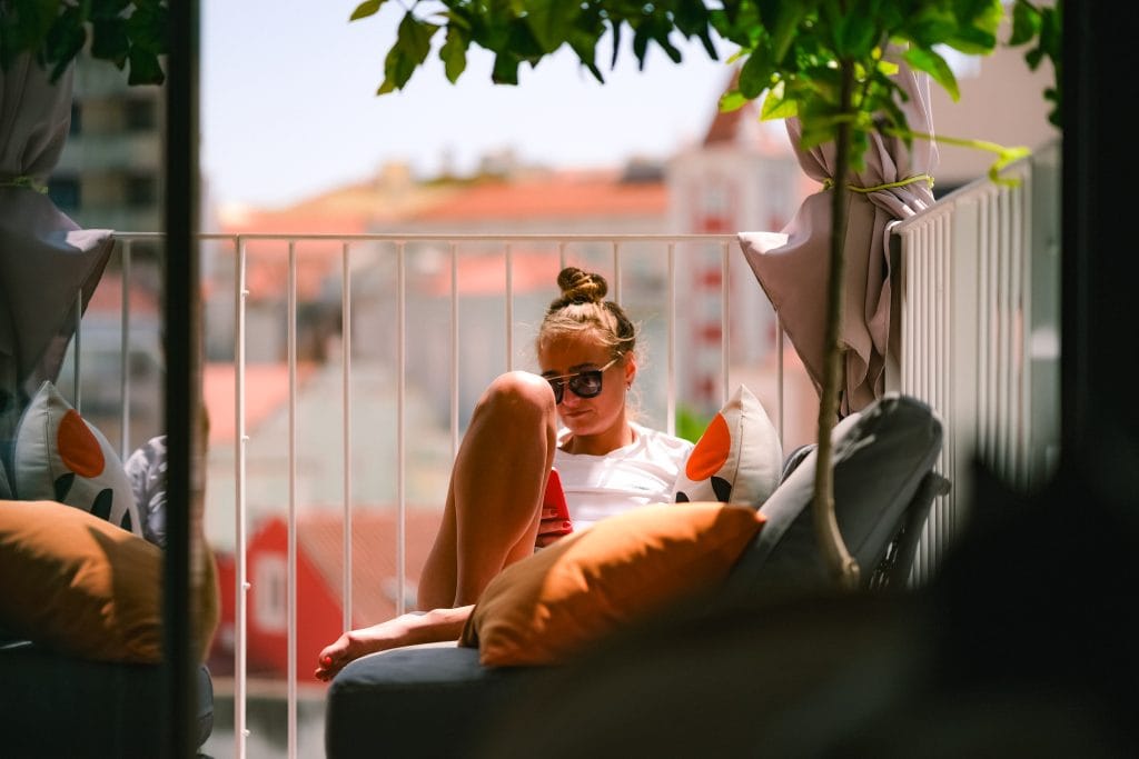 A woman wearing sunglasses relaxes on a balcony sofa, reading her phone in the sunlight while spending a month in Lisbon.