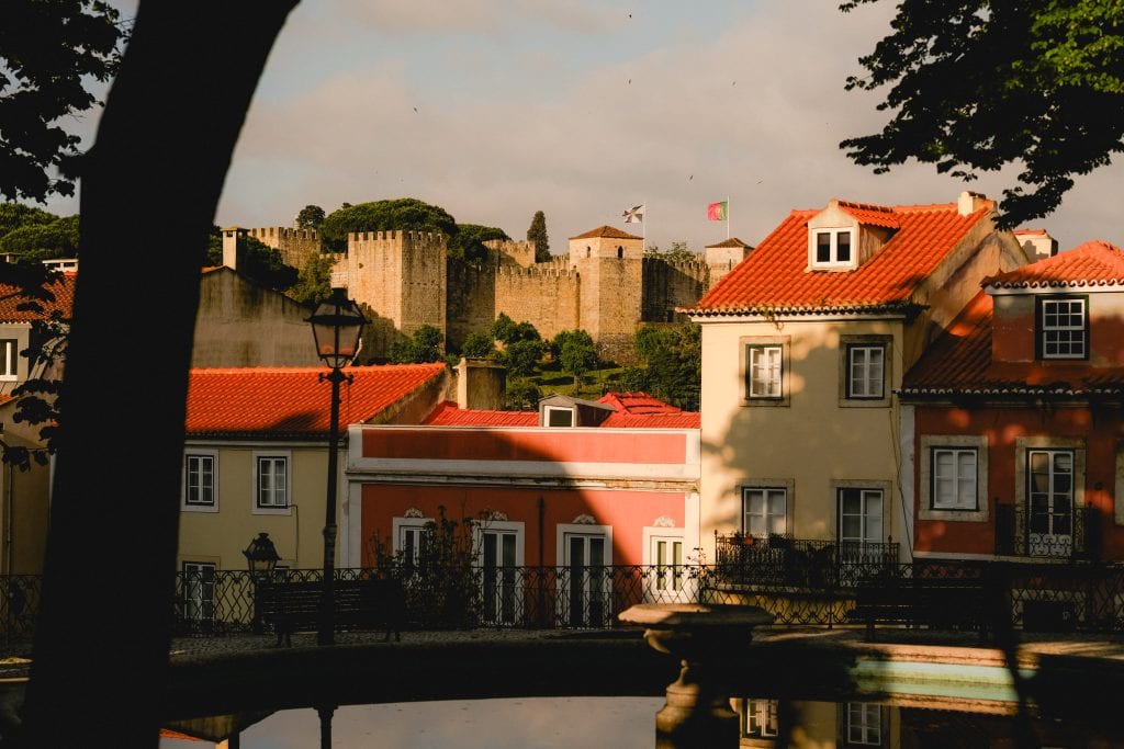 Colorful houses with red roofs, a lamp post, and a castle on a hill under a partly cloudy sky.