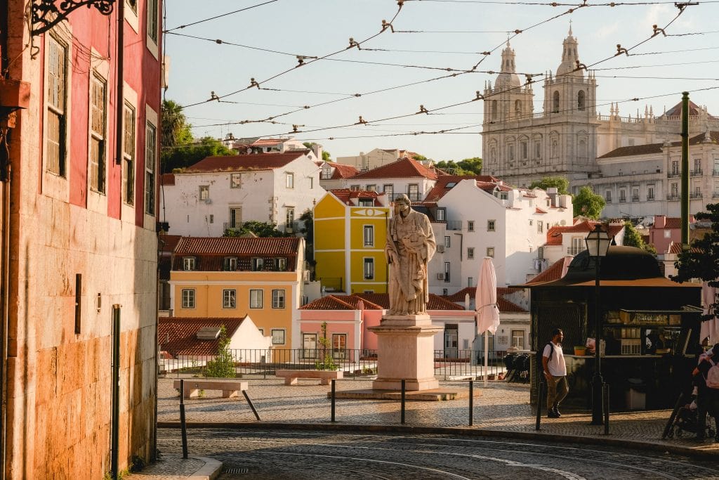 A statue in a sunny plaza in Lisbon with colorful buildings and a large church in the background.
