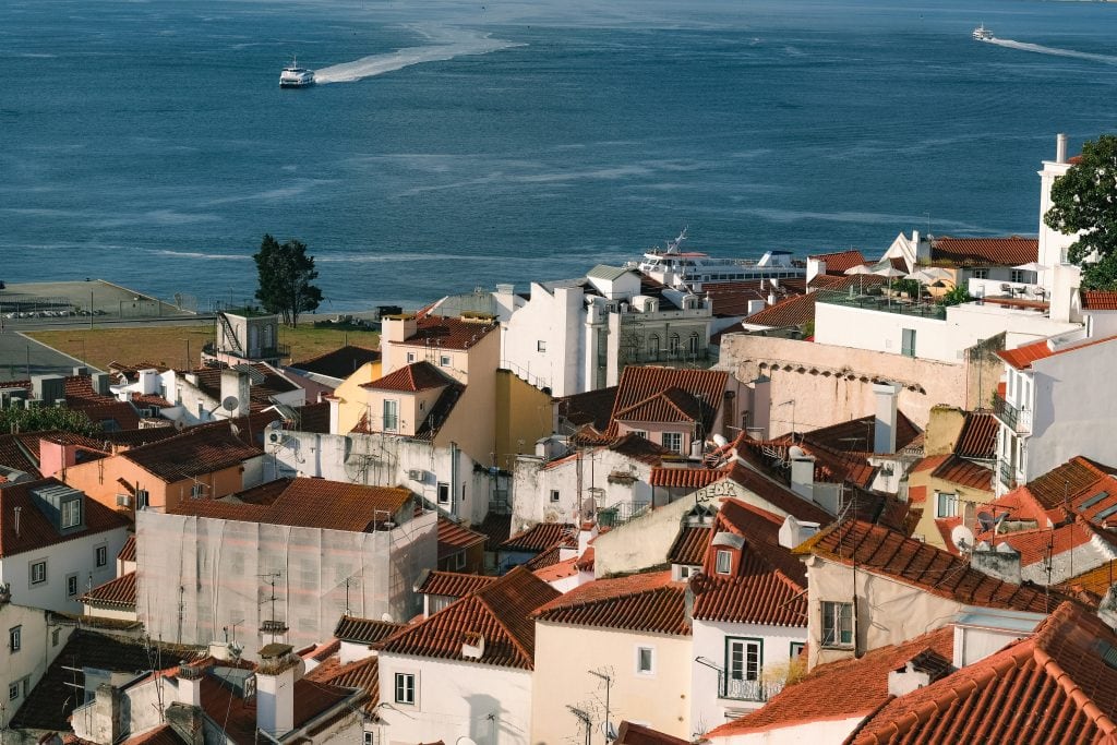 Residential buildings with white painted walls and terracotta roof slates cascade down a hill and meet a waterfront, busy with pleasure boats.