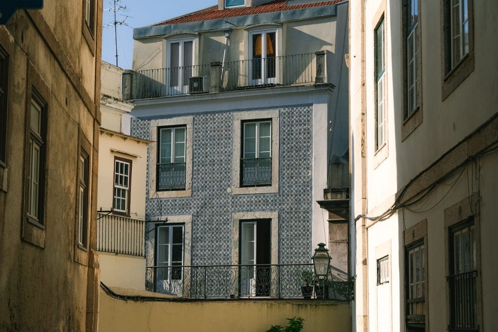 One of Lisbon's famous blue-tiled buildings with balconies is seen between two sunlit beige buildings in a narrow street.