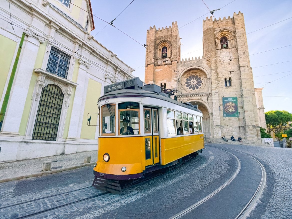 A yellow tram passes in front of a historic stone cathedral on a cobblestone street in Lisbon, Portugal.