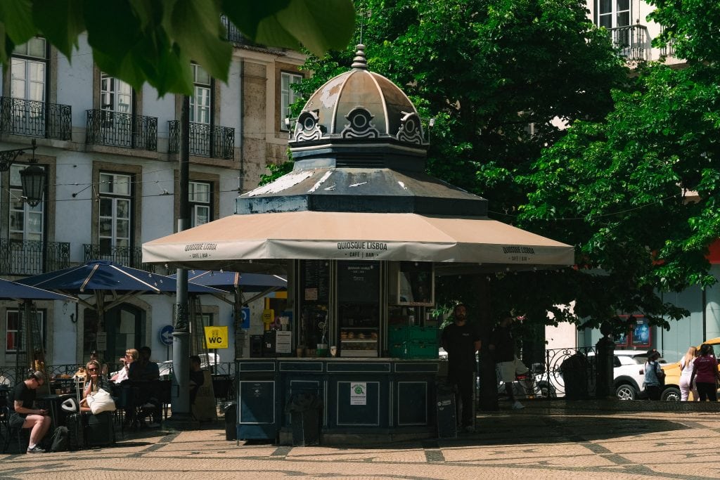 A small, ornate kiosk, Quiosque Lisboa, stands in a sunny plaza, surrounded by trees, people, and outdoor seating.