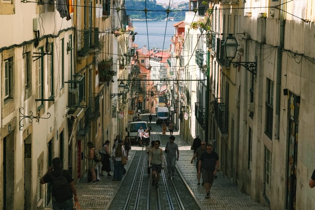 People walk up a hill between the tram tracks on a cobblestone street on a sunny day in Lisbon.