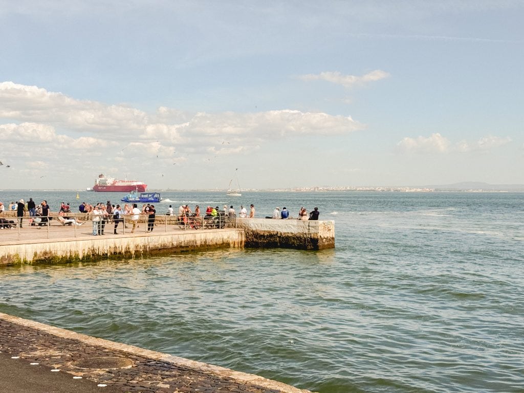 People stand on a pier by the sea, with a red ship and sailboats in the distance under a partly cloudy sky in Lisbon.