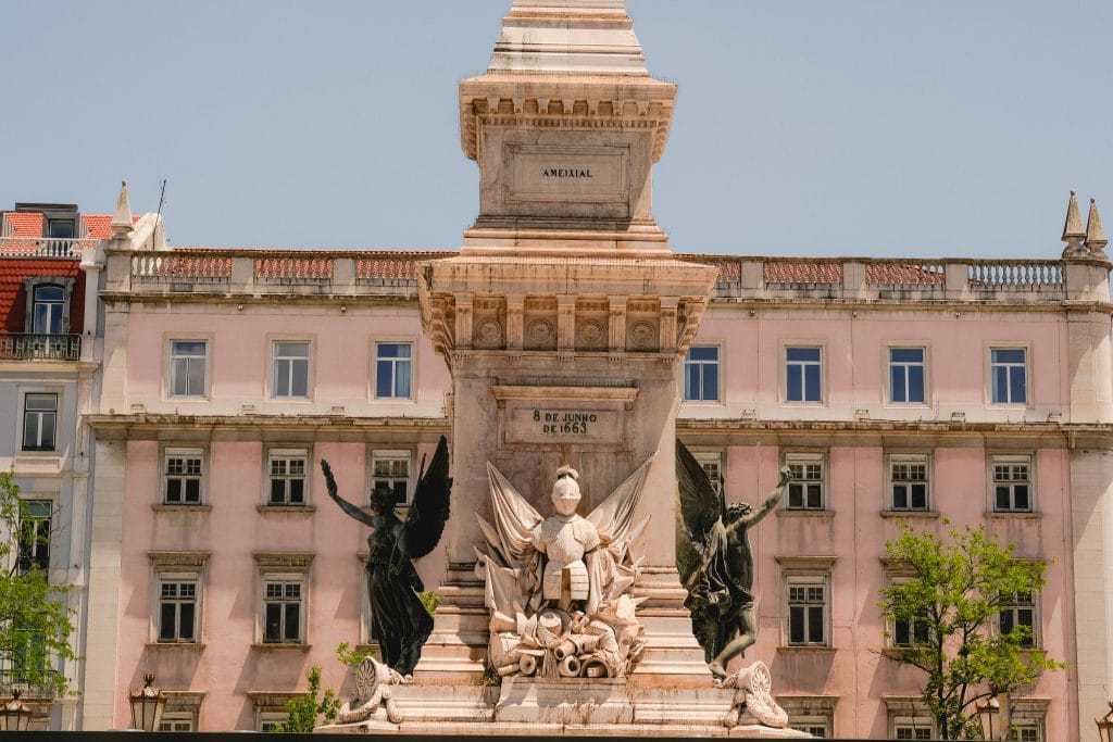 Stone monument with statues and angel figures in front of a pink building with windows and trees.