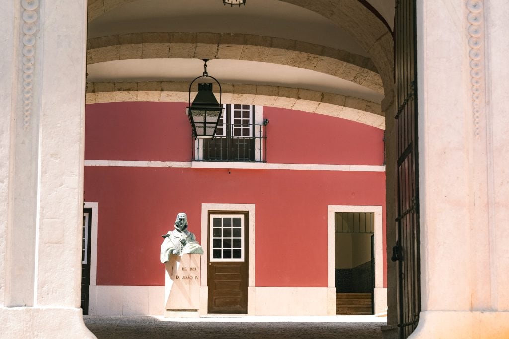 A statue stands in a sunlit courtyard with red walls, arched ceiling, and open doors.