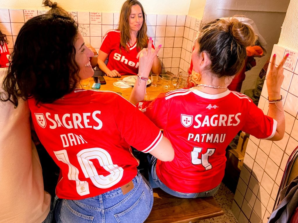 Three women in red Benfica jerseys sit at a table, smiling and making peace signs in a lively, tiled room.