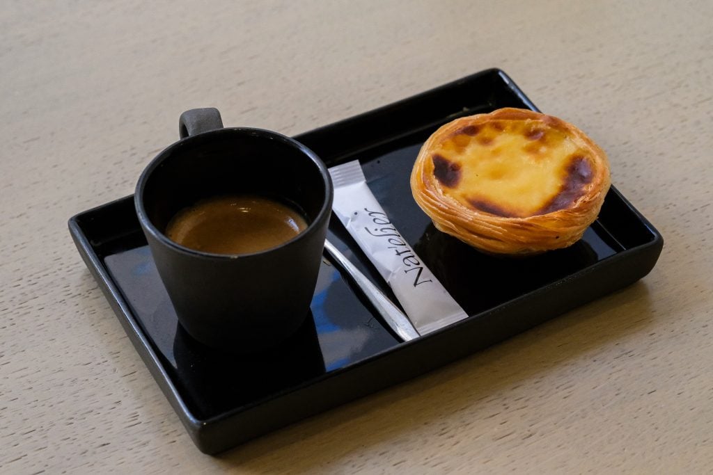 A pastel de nata on a black tray next to an espresso, with a sugar sachet branded 'Nat'elier'