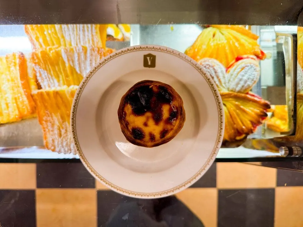 A pastel de nata on a decorative plate, displayed in a bakery with assorted pastries in the background.