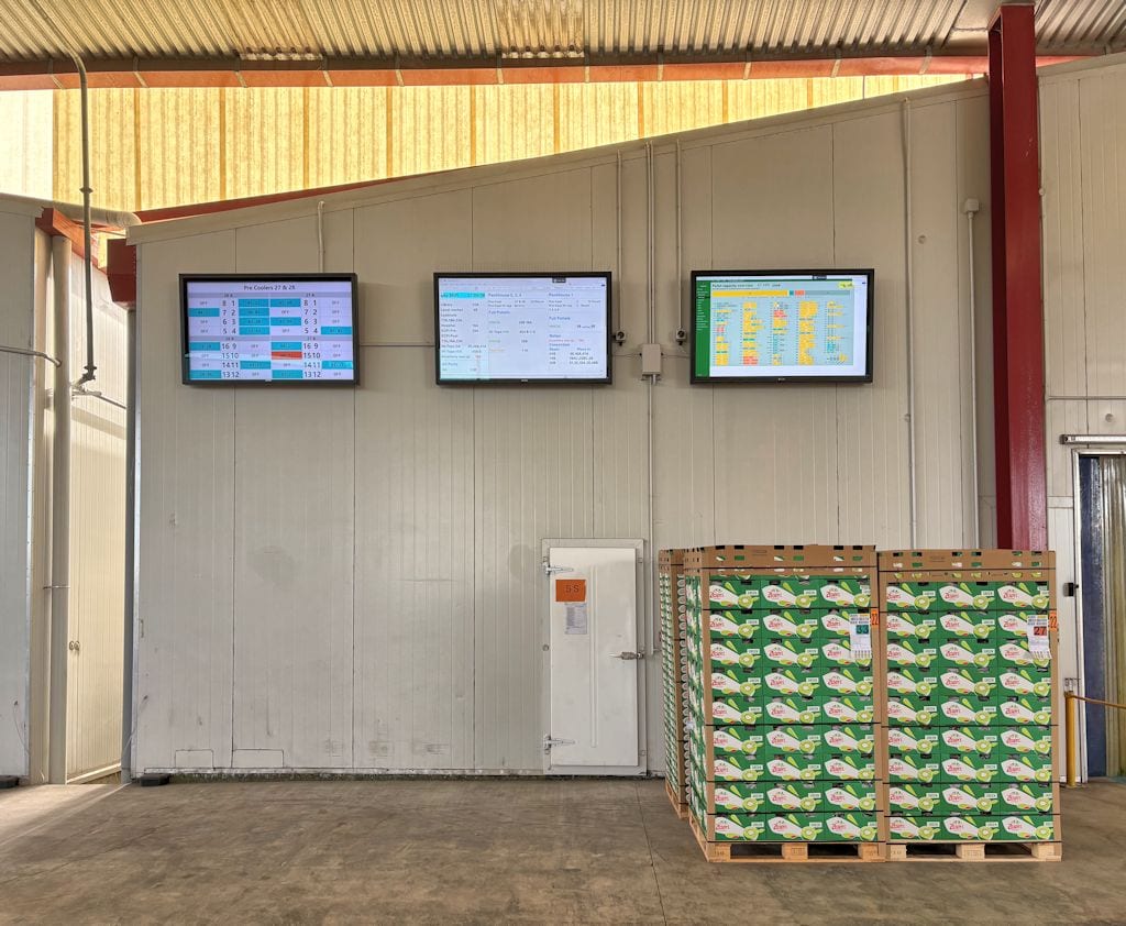 Warehouse interior with three monitors displaying schedules and data on a gray wall; pallets of green-and-white boxed goods to the right.
