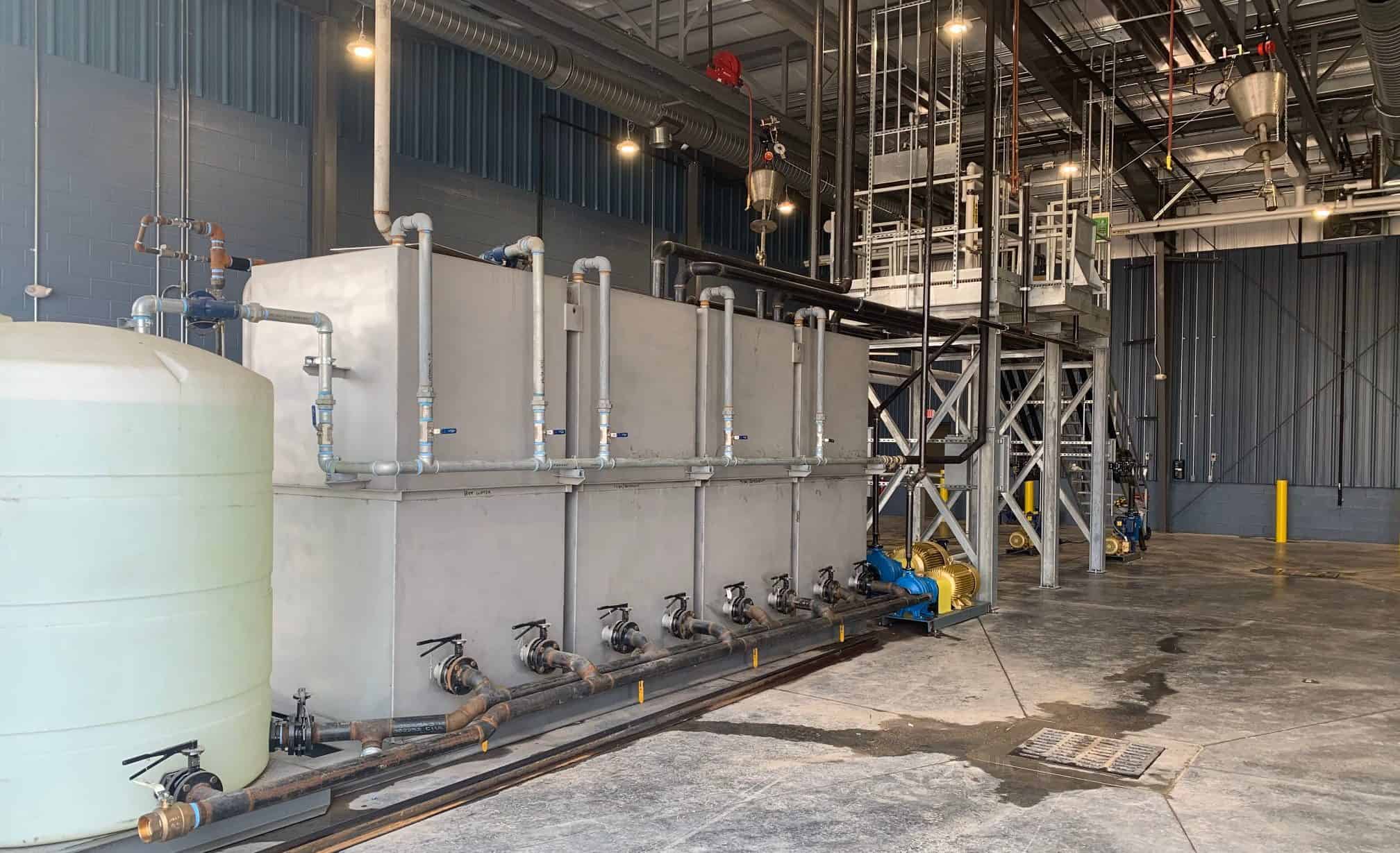 Industrial setting with large metal tanks and plumbing, housed in a warehouse with high ceilings and large doors.