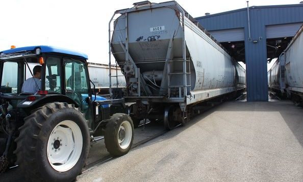 A tractor pulling a large white industrial tank between train cargo compartments in a warehouse area.