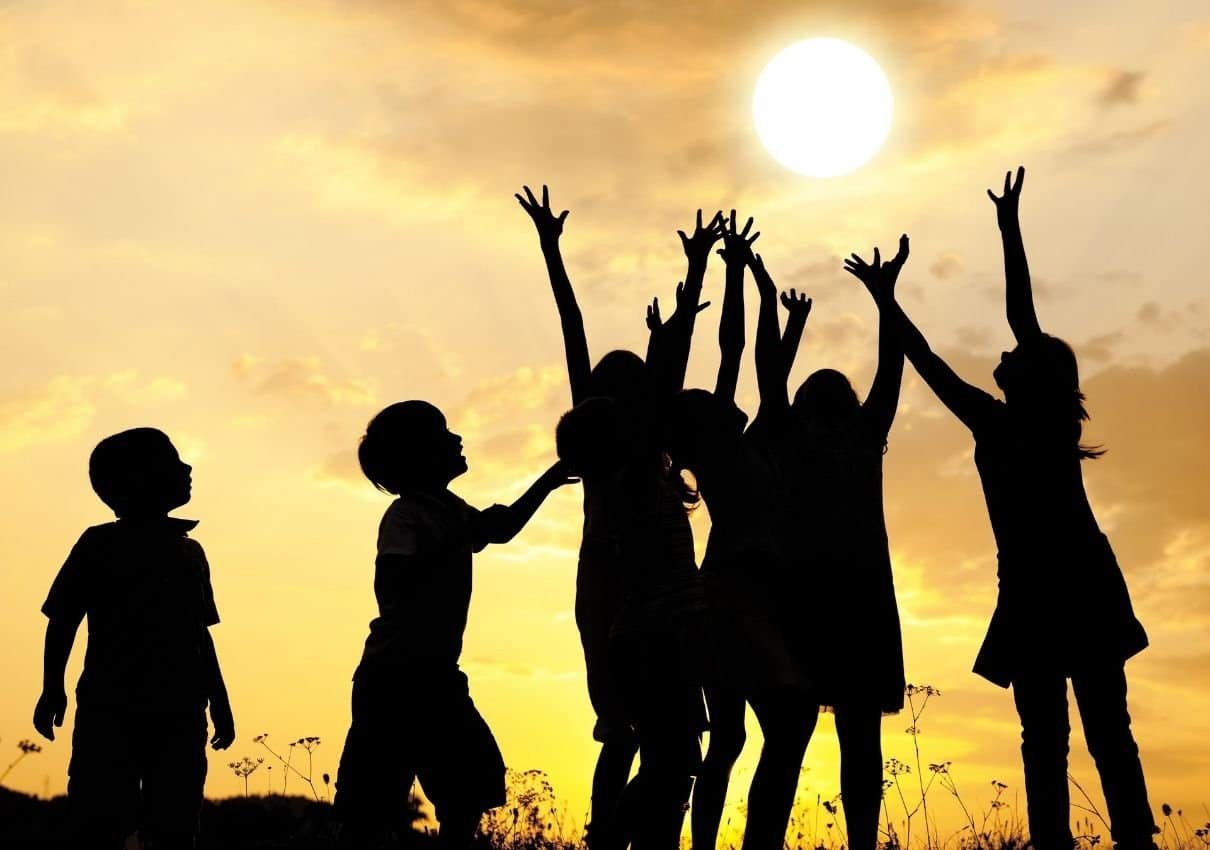 Silhouettes of children with arms raised towards a bright setting sun