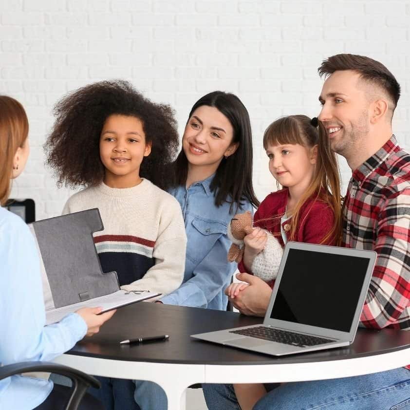 A professional speaking with a family of four, including two parents and two young girls, at a desk with a laptop and documents.