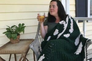 Woman relaxing with a drink on porch, wrapped in a green and white blanket.