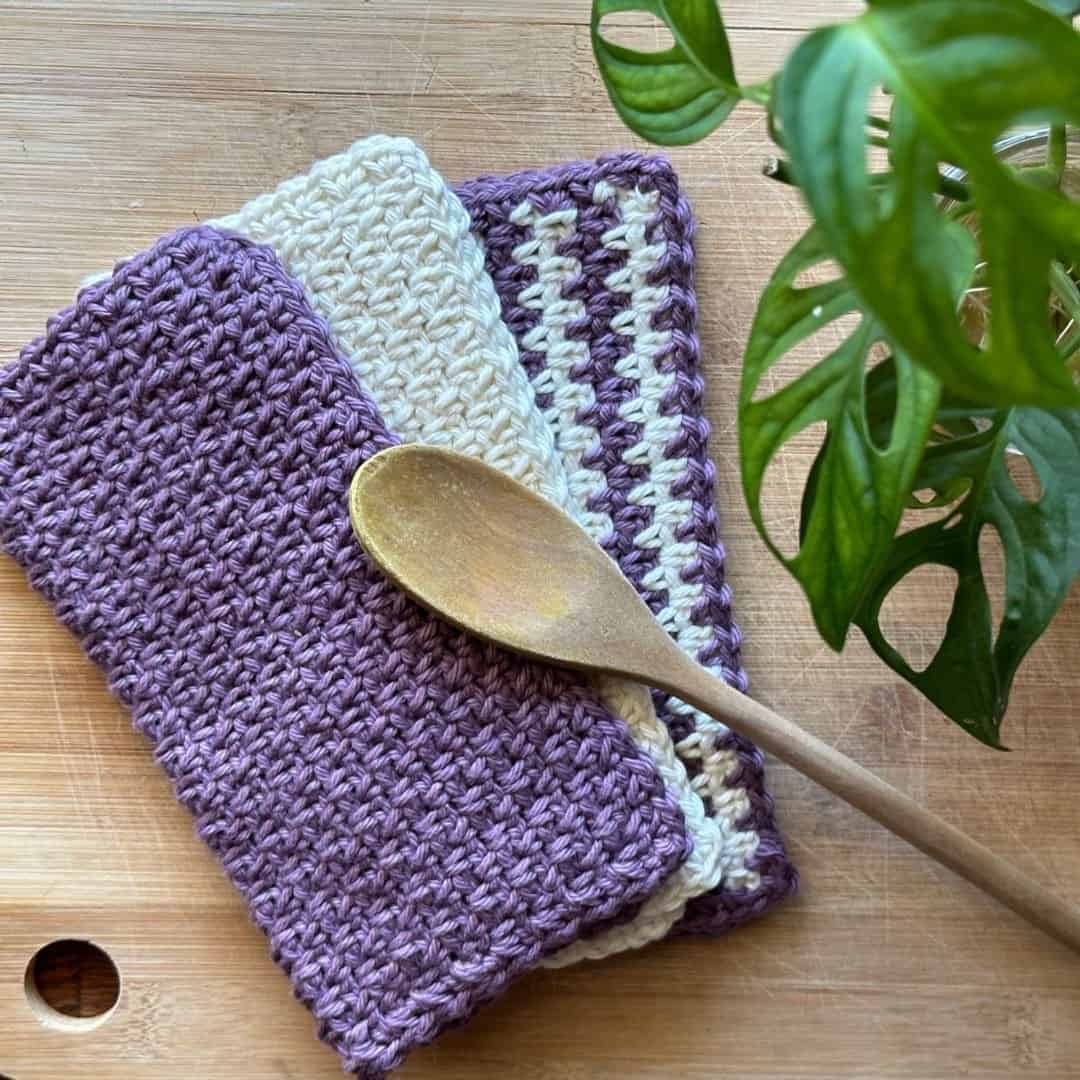Set of three handmade crochet washcloths in purple and white with moss stitch pattern.